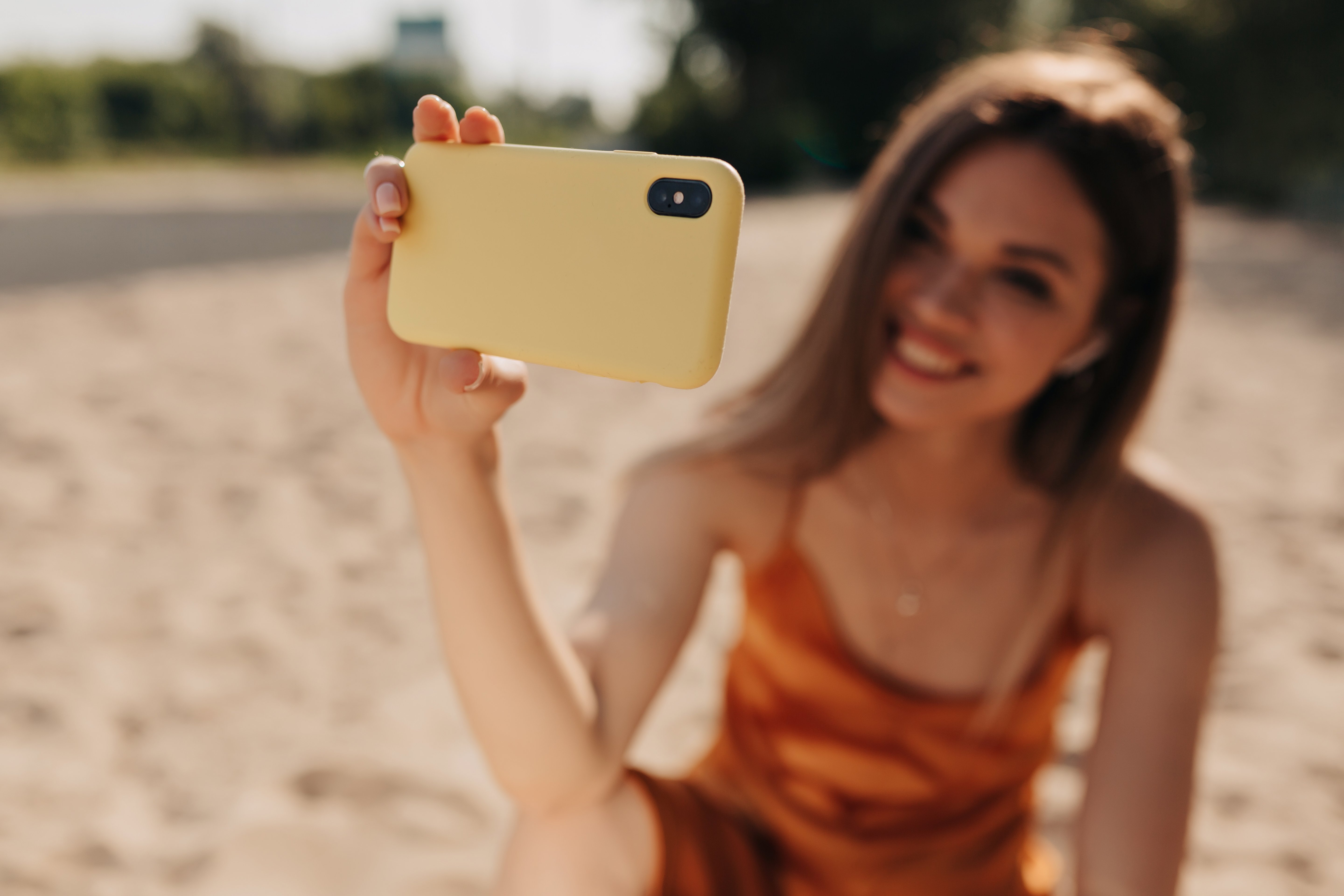 lovely-smiling-girl-with-dark-hairstyle-is-making-selfie-outdoors-sunshine-while-resting-beach-phone-focus-foreground-girl-background-out-focus